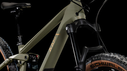 Nahaufnahme des CUBE Stereo ONE44 HPC Rookie dustyolive'n'gold 2025: modernes Mountainbike mit olivgrünem Carbonrahmen, schwarzer Federgabel, hellbraunen Schwalbe Nobby Nic-Reifen, auffälligen geometrischen Schläuchen und sichtbaren Stoßdämpfern.