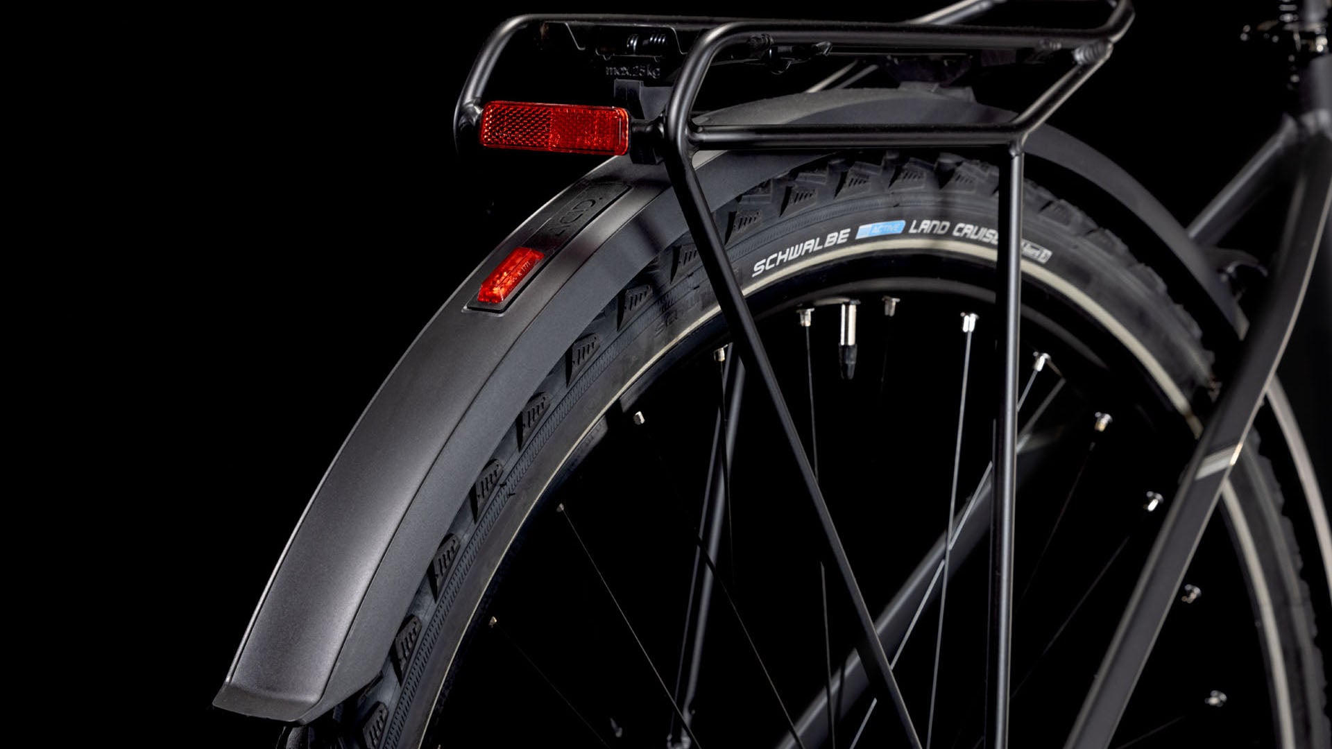 Nahaufnahme des Hinterrads eines CUBE Nature ONE Allroad black'n'black mit schwarzem Schutzblech, Gepäckträger, rotem Reflektor und Schwalbe Land Cruiser Reifen vor schwarzem Hintergrund. Shimano 1x8-Gang Schaltung Komponenten sind auch teilweise sichtbar.