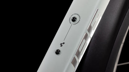 Nahaufnahme eines CUBE Nature EXC Allroad seasalt'n'pyrite 2025, weißer Rahmen mit Metallic-Logo, schwarzer Schraube und Aufschrift "Designed and Engineered in Germany". Im Hintergrund ist ein Teil eines Rades mit schwarzem Reifen zu sehen.