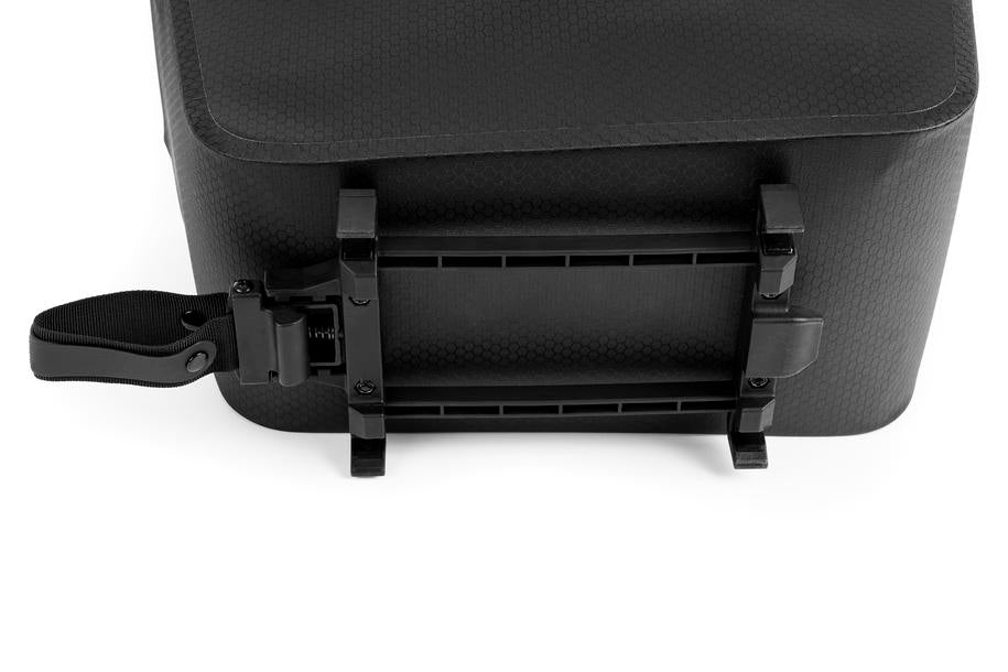 Eine Nahaufnahme zeigt die Unterseite einer schwarzen, rechteckigen Tasche mit einer Kunststoffhalterung, die mit dem ACID RILink Adapter Pro kompatibel ist, und einem verstellbaren Riemen zur Sicherung der Ladung auf einer Seite.
