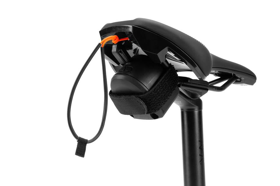 Ein Cube ACID Zugsystem MOUNTAIN EXPLORER schwarzer Fahrradsattel mit einer kleinen schwarzen Tasche, die mit einem Riemen darunter befestigt ist, mit einem selbstaufrollenden Mechanismus und einer orangefarbenen Zuglasche mit schwarzer Schlaufe, dargestellt auf einem weißen Hintergrund.
