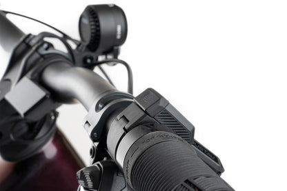 Nahaufnahme eines Fahrradlenkers mit strukturiertem Gummigriff, ACID E-Bike Frontlicht PRO-E 200 High Beam X-Connect schwarz mit StVZO-Zertifizierung und Steuerschalter von ACID vor weißem Hintergrund.