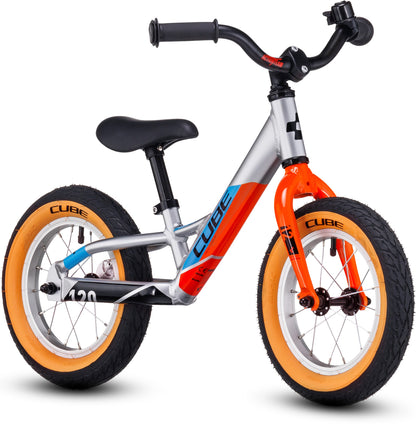 Das CUBE Cubie 120 walk actionteam Laufrad von Cube ist ein leichtes Kinder-Balancebike mit silbernem, orangefarbenem und blauem Aluminiumrahmen, schwarzem Sitz und Lenker, beigen Reifen und CUBE-Branding auf Rahmen und Rädern.
