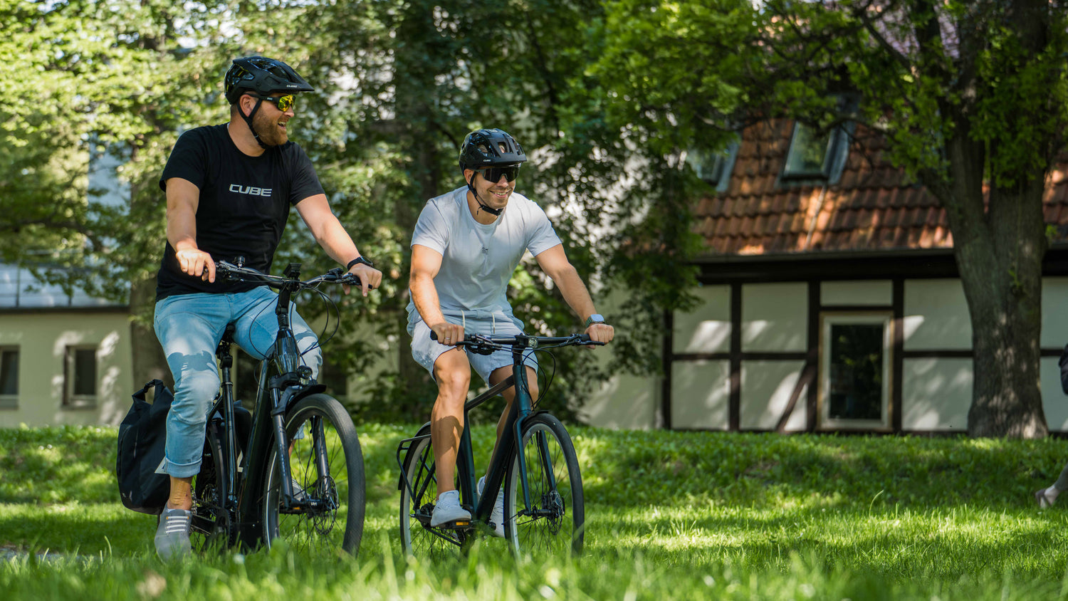 Zwei Personen mit Helmen fahren mit CUBE-Fahrrädern auf einem Grasweg in einem sonnigen Park. Lächelnd genießen sie die Natur. Im Hintergrund sind Bäume und ein Gebäude mit Ziegeldach zu sehen – eine perfekte Kulisse für Radsportfans in Niedersachsen.