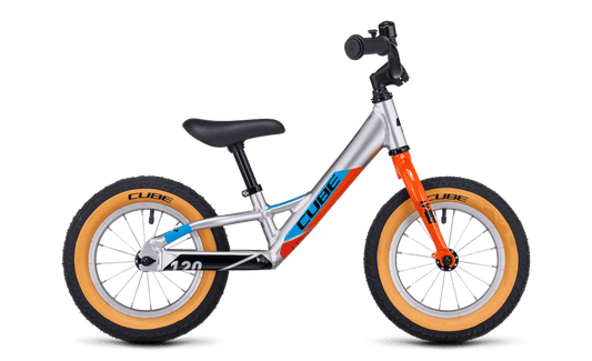 Das Cube Cubie 120 walk actionteam Laufrad ist ein leichtes Aluminium-Balancebike in Silber mit orangefarbener Gabel, schwarzem Sitz, blauem CUBE Branding und beigen Reifen. Es hat keine Pedale oder Stützräder und ist auf einem schlichten weißen Hintergrund abgebildet.