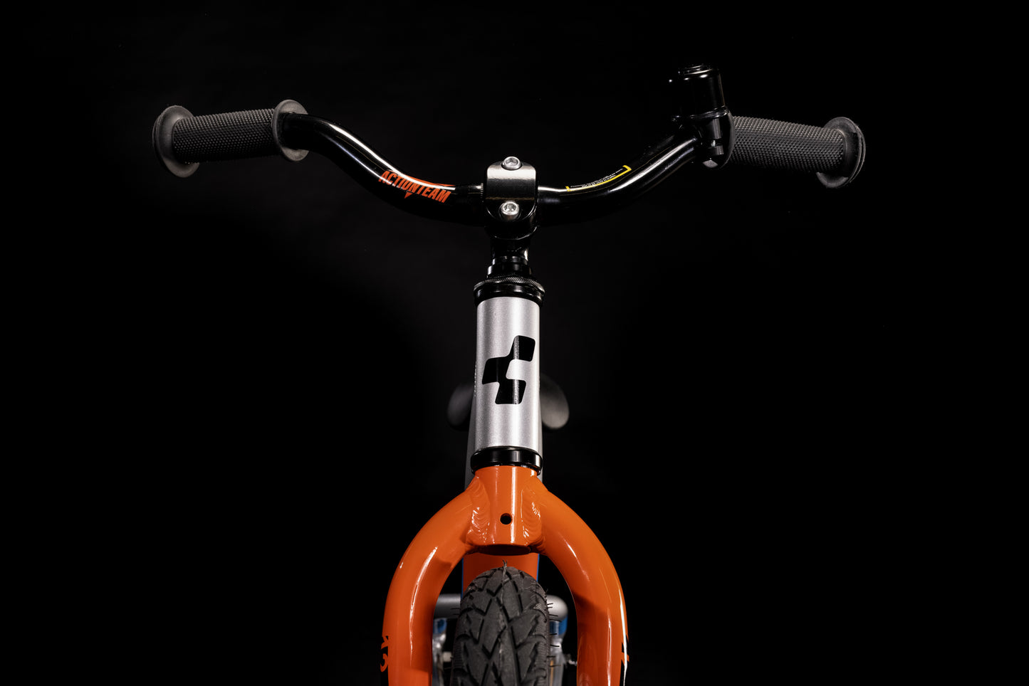 Nahaufnahme der Vorderseite des Cube CUBE Cubie 120 walk actionteam Laufrad, mit orangefarbener Gabel, dickem schwarzem Vorderreifen und breitem Lenker. Dieses leichte Aluminium-Fahrrad hebt sich wunderbar von einem schwarzen Hintergrund ab.