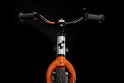 Nahaufnahme der Vorderseite des Cube CUBE Cubie 120 walk actionteam Laufrad, mit orangefarbener Gabel, dickem schwarzem Vorderreifen und breitem Lenker. Dieses leichte Aluminium-Fahrrad hebt sich wunderbar von einem schwarzen Hintergrund ab.